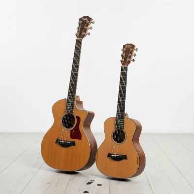 A side-by-side comparison of a Taylor GS Mini Acoustic Guitar and a Martin LX1 Little Martin Acoustic Guitar on a clean, light-colored wooden table, showcasing their differences in body shape, size, and detailing in a well-lit, professional product photography style.